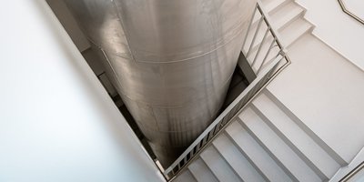 Eine moderne Treppe führt hinunter zu einem großen, silbernen Zylinder, der in einem hellen Raum steht.