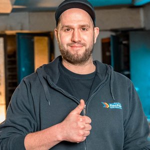 Ein Mann mit Bart und Mütze lächelt und zeigt ein Daumen-hoch-Zeichen in einer Werkstatt mit blauen Lichtern im Hintergrund.