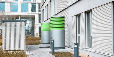 Zwei große, grüne Zylinder stehen neben einem modernen Gebäude, umgeben von einem gepflegten Garten und einem gepflasterten Weg.