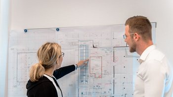 Eine Frau zeigt auf einen detaillierten Bauplan, während ein Mann aufmerksam zuhört und die Informationen aufnimmt.