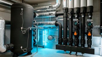 Ein moderner Technikraum mit einem großen schwarzen Wassertank und mehreren Rohren, die an der Wand verlaufen.
