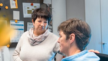 Eine Frau mit einem Schal spricht mit einer anderen Frau, die in einem Stuhl sitzt, in einem hellen Büro.
