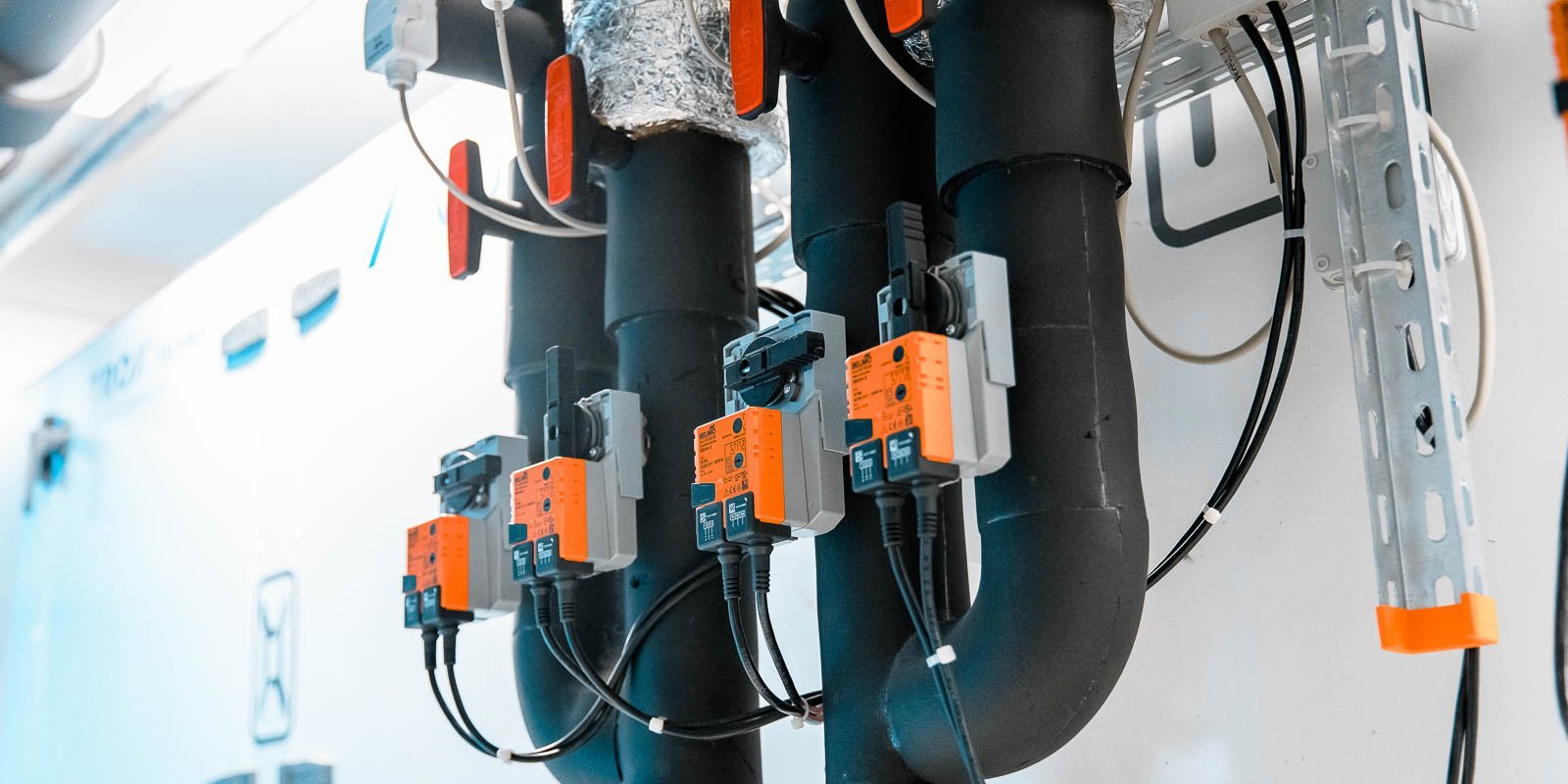 Eine Reihe von schwarzen Rohren mit orangefarbenen Sensoren und Kabeln, die an einer Wand montiert sind, in einem technischen Raum.