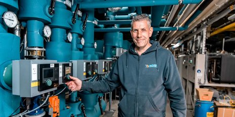 Ein Techniker steht lächelnd vor einer komplexen Anlagentechnik mit blauen Rohren und Steuergeräten.