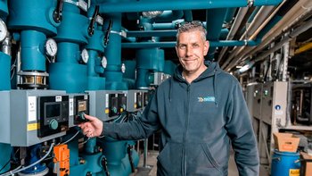Ein Techniker steht lächelnd vor einer komplexen Anlagentechnik mit blauen Rohren und Steuergeräten.