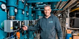 Ein Techniker steht lächelnd vor einer komplexen Anlagentechnik mit blauen Rohren und Steuergeräten.