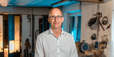 Ein lächelnder Mann in einem hellen Hemd steht in einer Werkstatt mit Werkzeugen und Maschinen im Hintergrund.