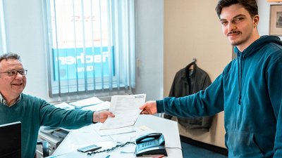 Ein junger Mann übergibt ein Dokument an einen lächelnden älteren Mann in einem Büro mit vielen Unterlagen.