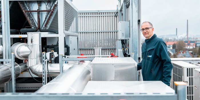 Ein Techniker steht auf einem Dach neben großen, silbernen Lüftungsrohren und schaut lächelnd in die Kamera.
