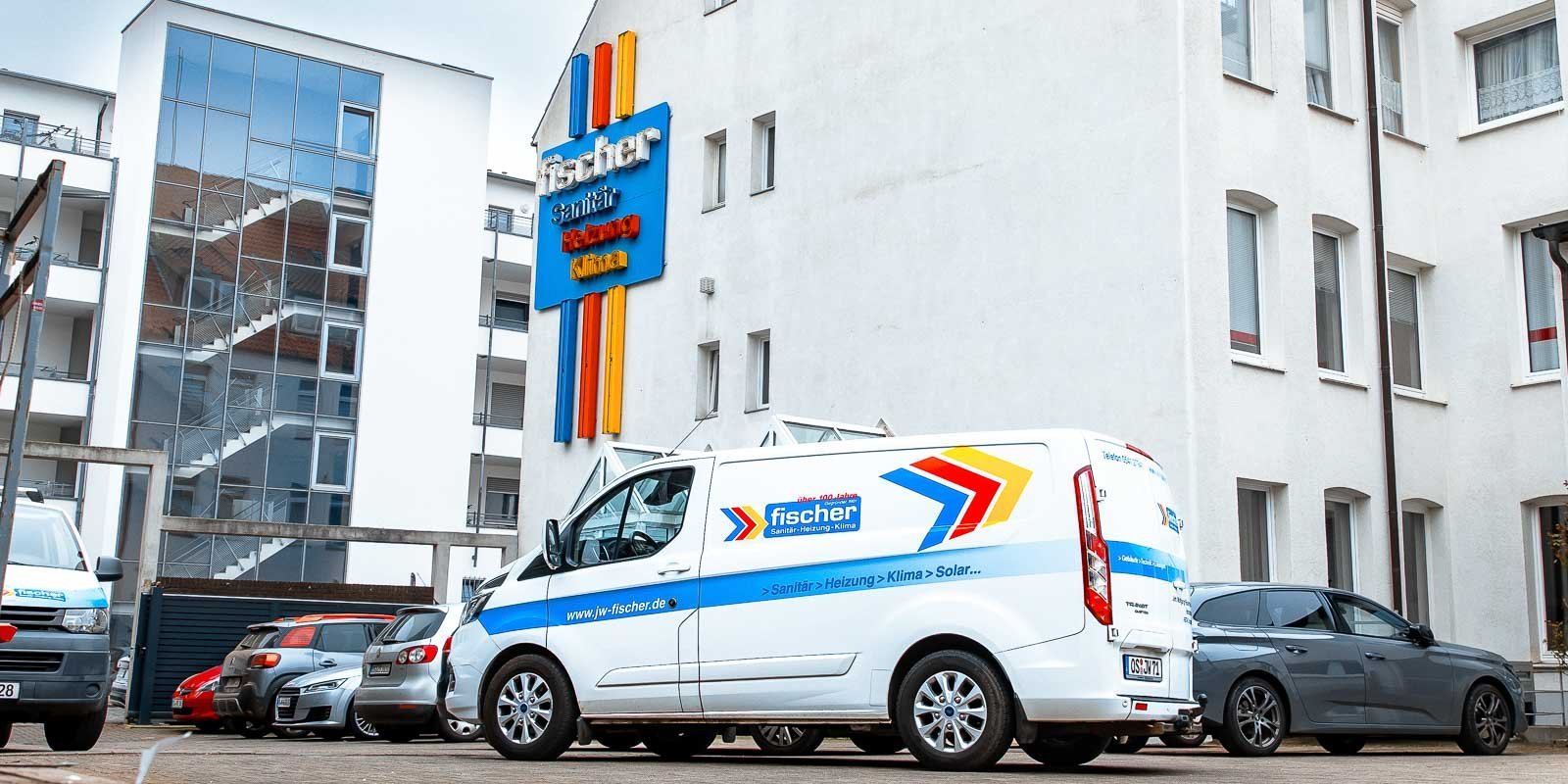 Ein weißer Lieferwagen mit dem Logo von Fischer steht auf einem Parkplatz vor einem modernen Gebäude mit großen Fenstern.