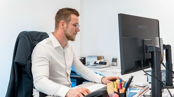 Ein Mann in einem weißen Hemd sitzt konzentriert an einem Schreibtisch und arbeitet an einem Computerbildschirm.