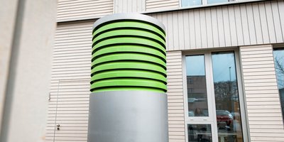 Ein modernes, zylindrisches Objekt mit grünen Rillen steht vor einem Gebäude mit Holzverkleidung und großen Fenstern.