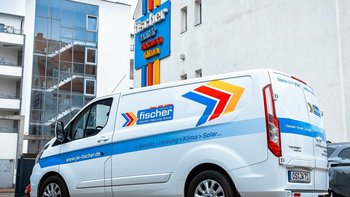 Ein Lieferwagen mit dem Logo von Fischer steht vor einem modernen Gebäude mit großen Fenstern und einer Treppe im Hintergrund.