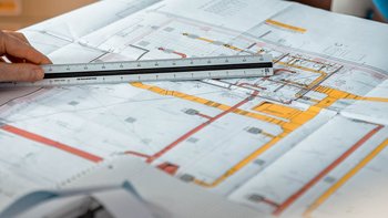 Eine Hand hält ein Lineal über einen detaillierten technischen Plan mit farbigen Markierungen und verschiedenen Linien.
