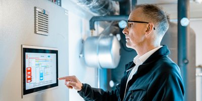 Ein Mann in einem schwarzen Jackett betrachtet konzentriert ein digitales Steuerungspanel in einem technischen Raum.