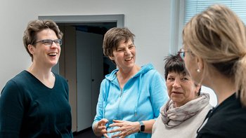 Vier Frauen stehen in einem hellen Raum und lachen miteinander, während sie sich angeregt unterhalten.