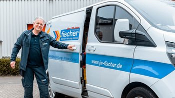 Ein Handwerker in Arbeitskleidung steht lächelnd neben einem weißen Lieferwagen mit dem Logo von fischer Sanitär-Heizung-Klima.