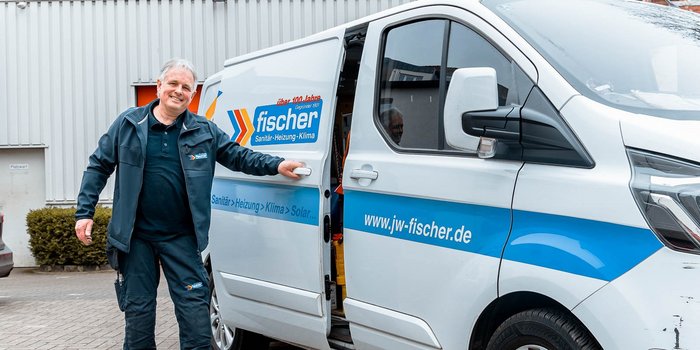 Ein Handwerker in Arbeitskleidung steht lächelnd neben einem weißen Lieferwagen mit dem Logo von fischer Sanitär-Heizung-Klima.