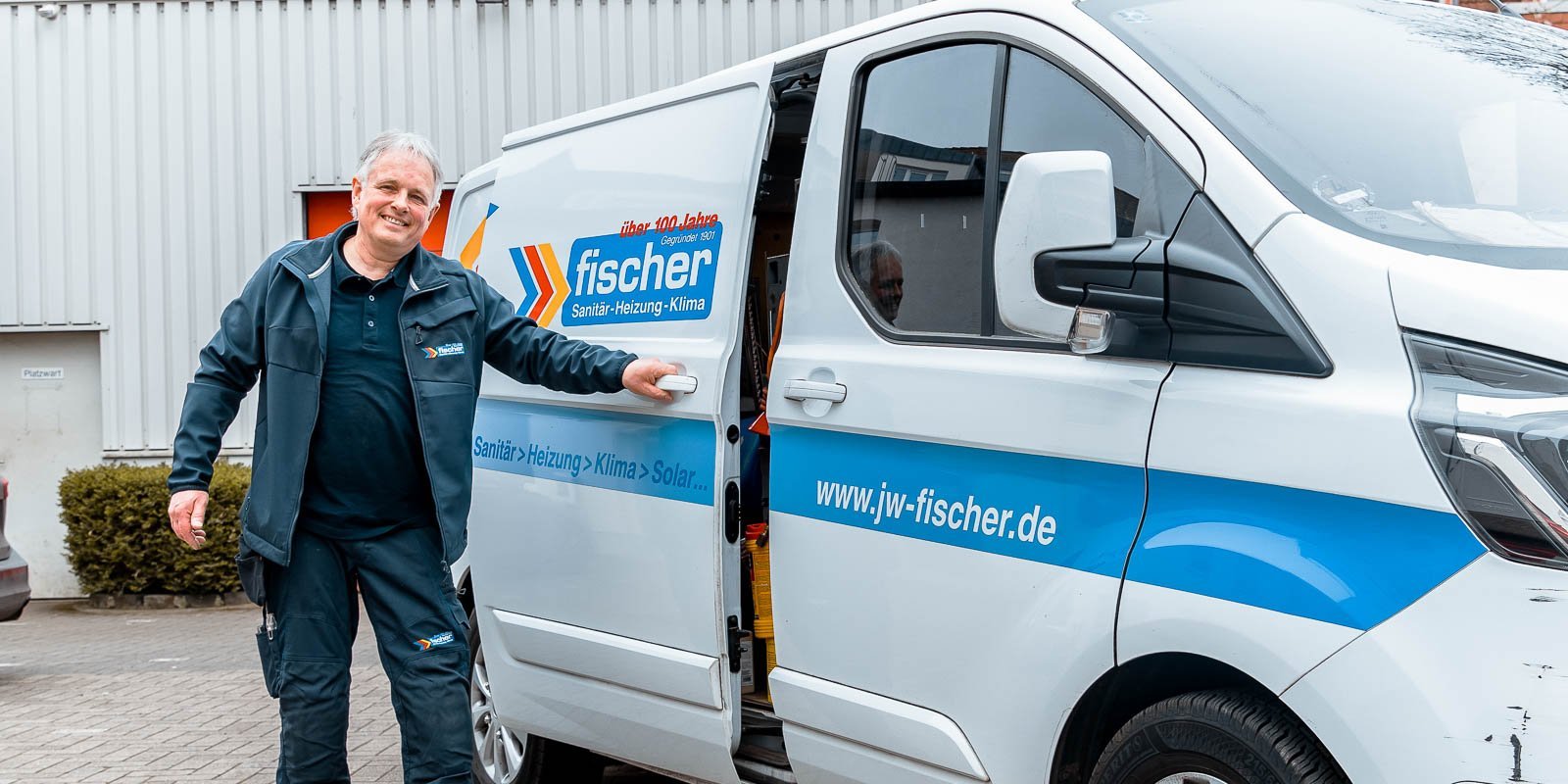 Ein Handwerker in Arbeitskleidung steht lächelnd neben einem weißen Lieferwagen mit dem Logo von fischer Sanitär-Heizung-Klima.