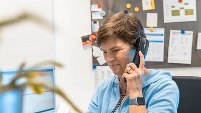 Eine Frau mit kurzem Haar lächelt, während sie am Telefon spricht, umgeben von einem organisierten Büro mit Notizen.