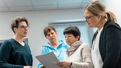 Vier Frauen stehen in einem hellen Raum und diskutieren angeregt über ein Dokument, das eine von ihnen hält.